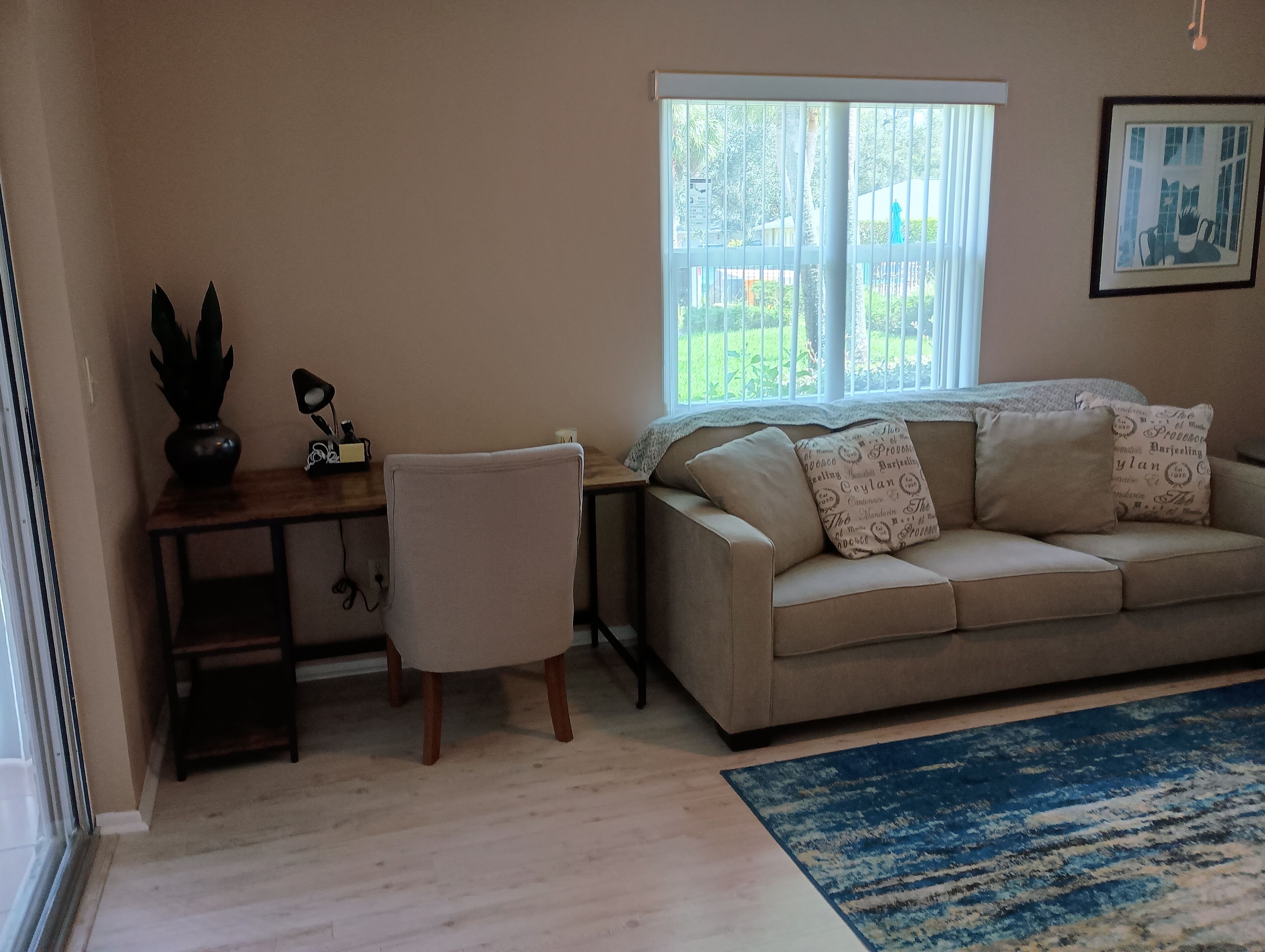 A living room with a beige sofa, a wooden desk, a chair, a blue rug, and a window with a view of greenery.