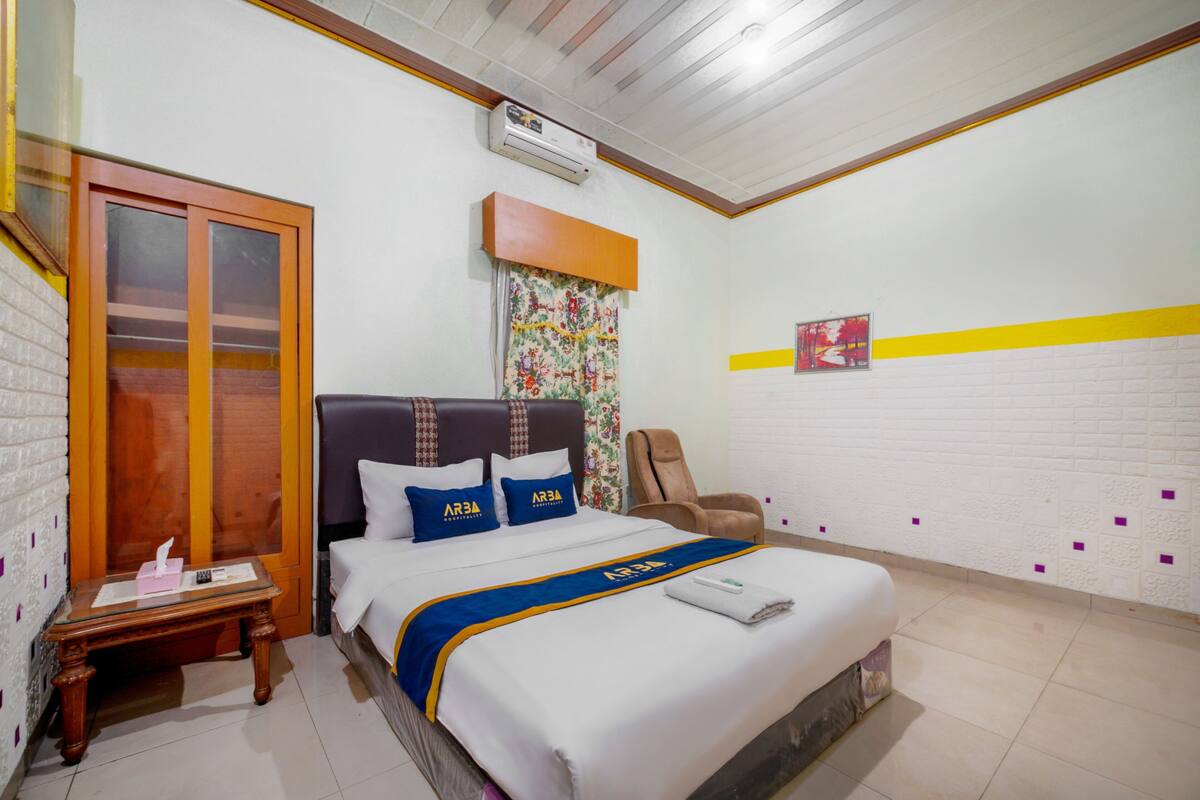 A hotel room with a bed, a chair, a table, and a window with blinds.