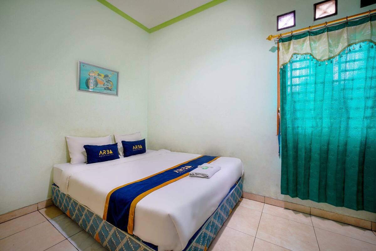 A neatly made bed with a blue and white bedspread, a blue pillow, and a green curtain on the window.