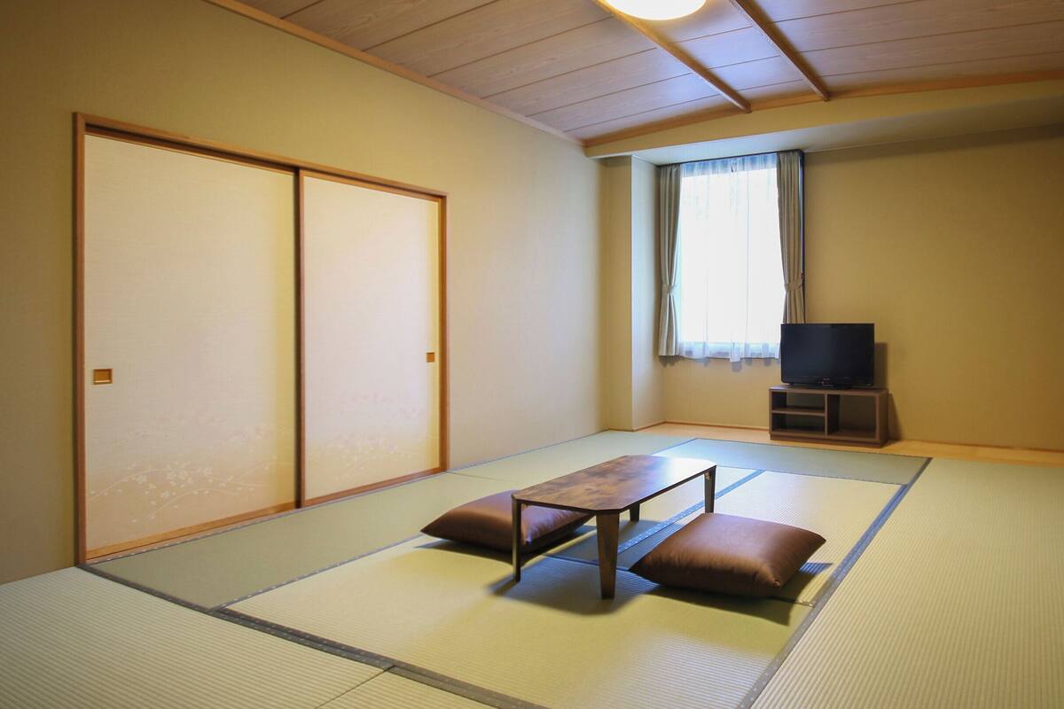 Una stanza tradizionale giapponese con pavimenti in tatami, un tavolino con cuscini, un televisore su un supporto e porte scorrevoli.