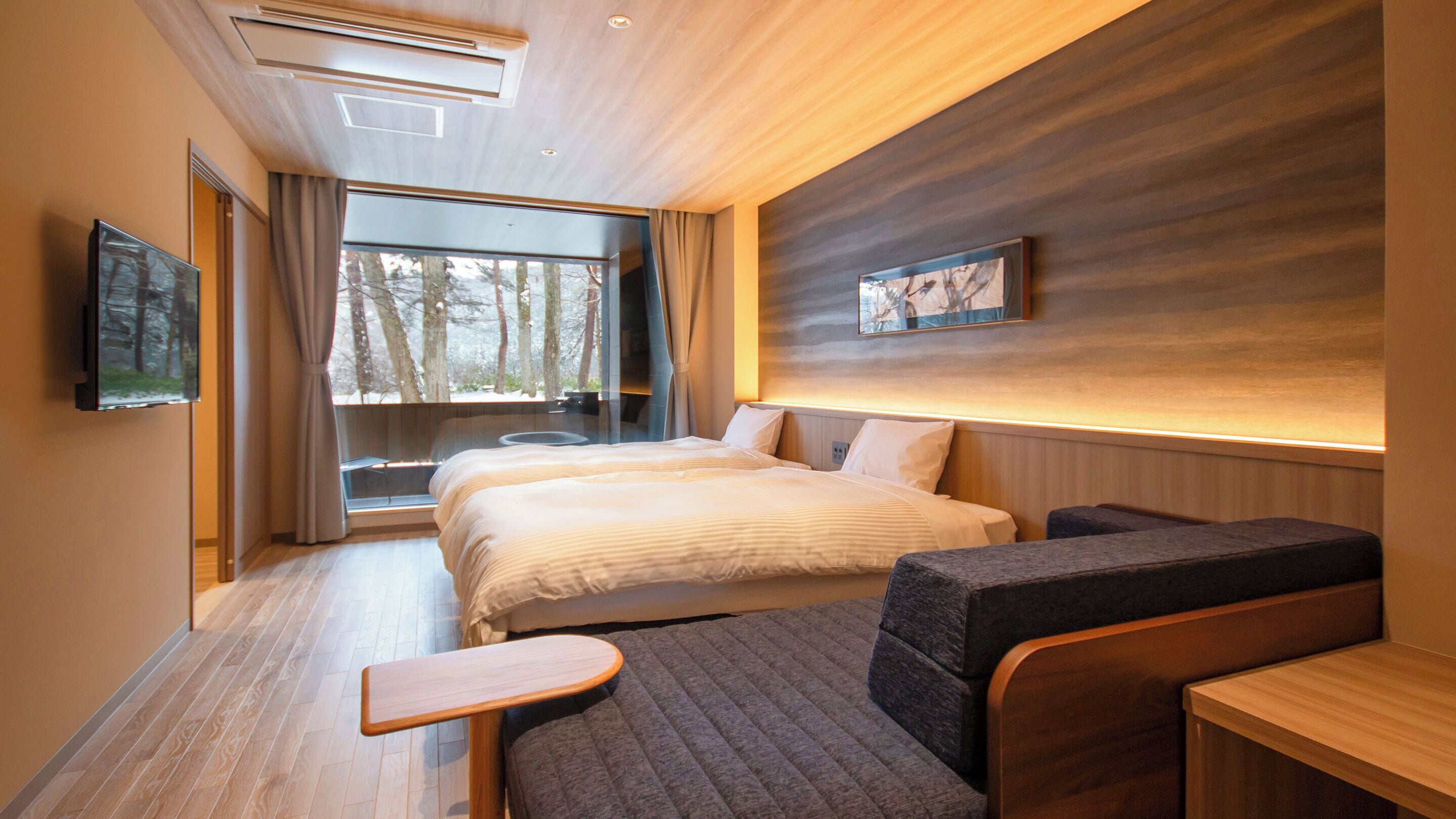 A modern hotel room with a large bed, a sofa, a small table, and a TV mounted on the wall.