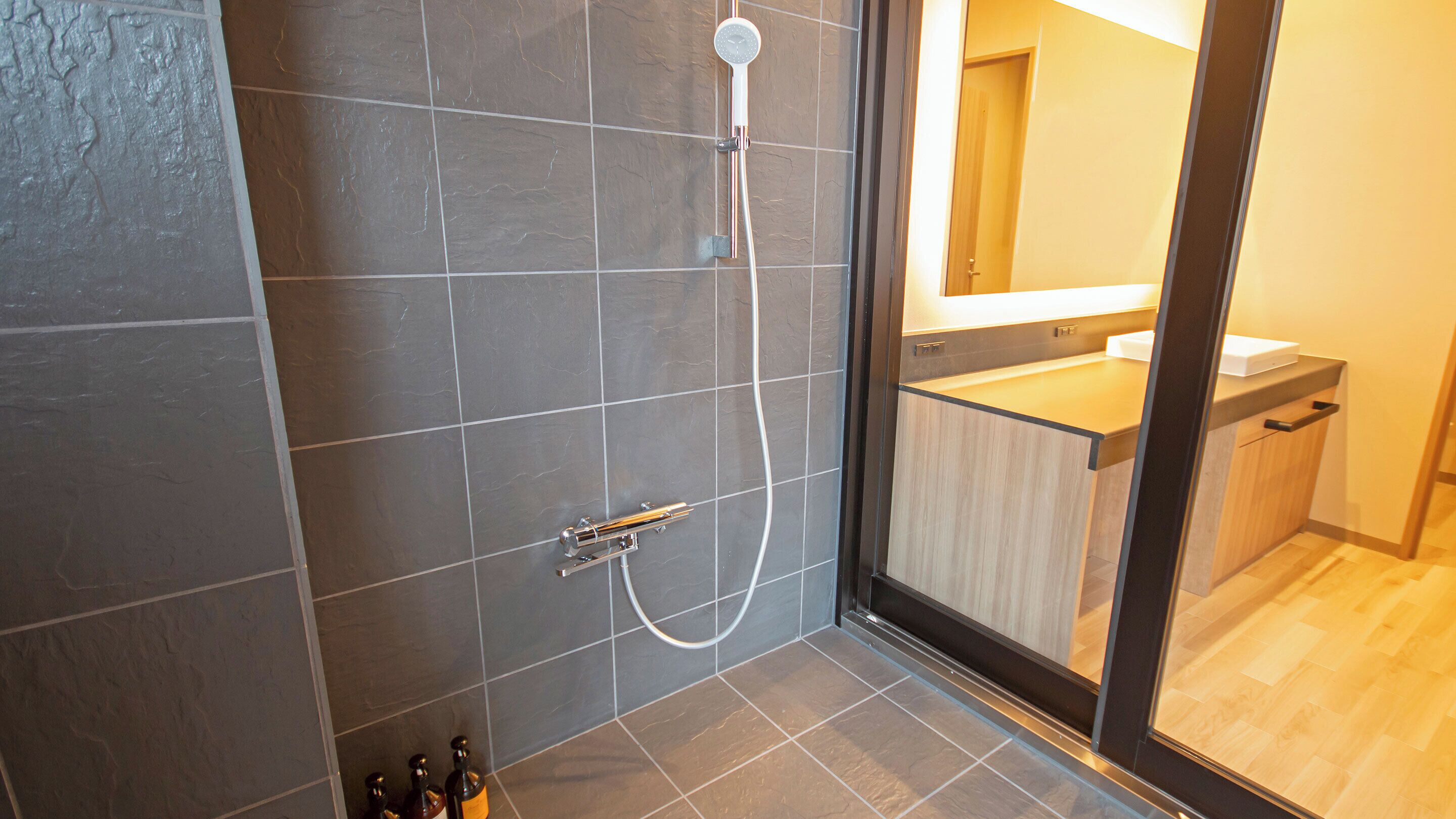 A modern bathroom with a glass shower enclosure, dark tiles, and a wooden vanity with a mirror.