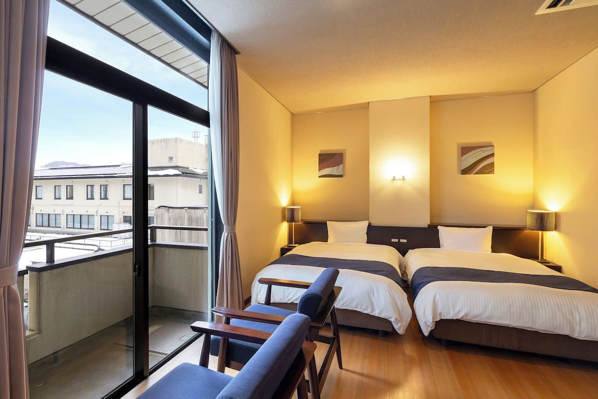 Camera d'albergo con due letti, un balcone con tende e vista sugli edifici.