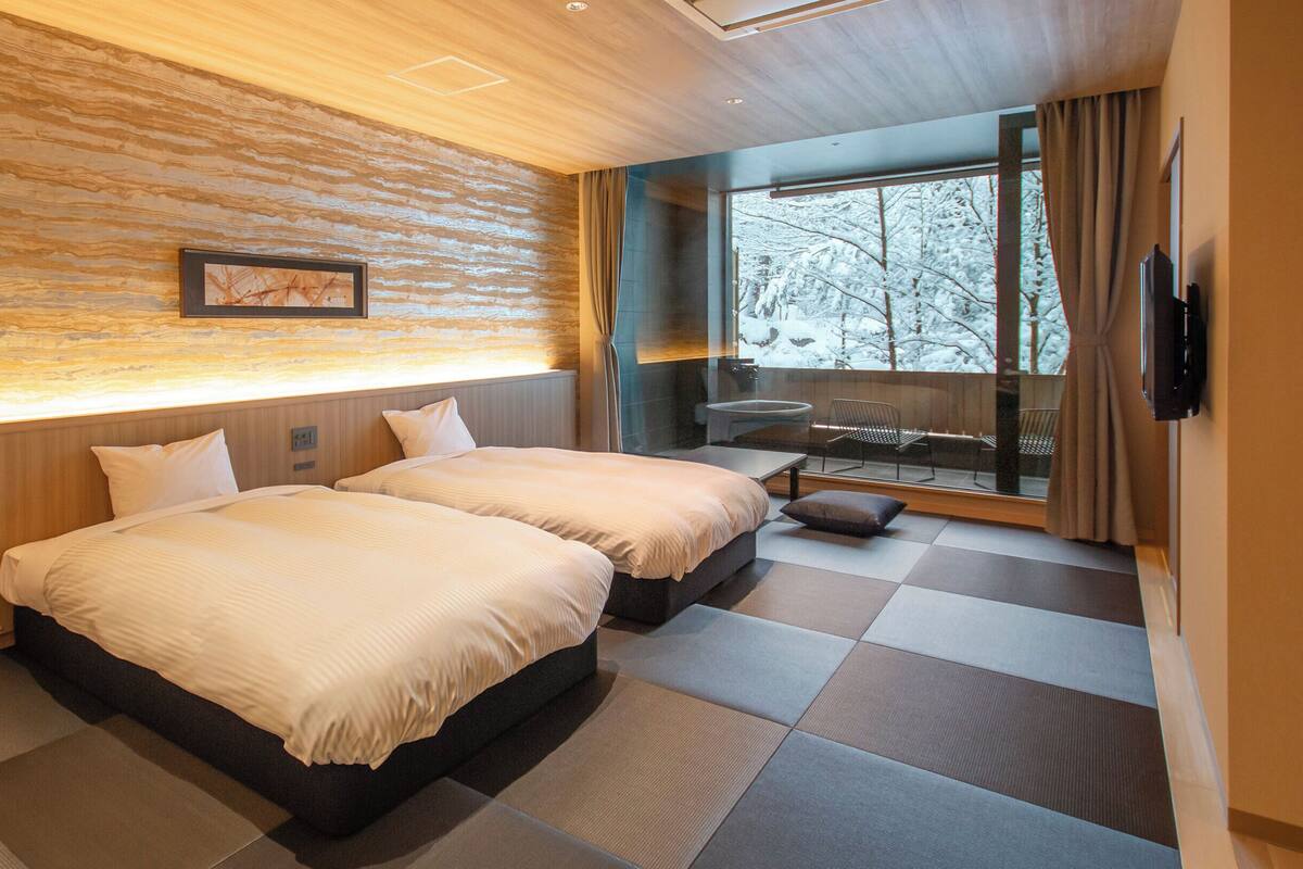 Una camera d'albergo moderna con due letti, un'ampia finestra che si affaccia su alberi innevati e un'area salotto con una sedia e un tavolino.