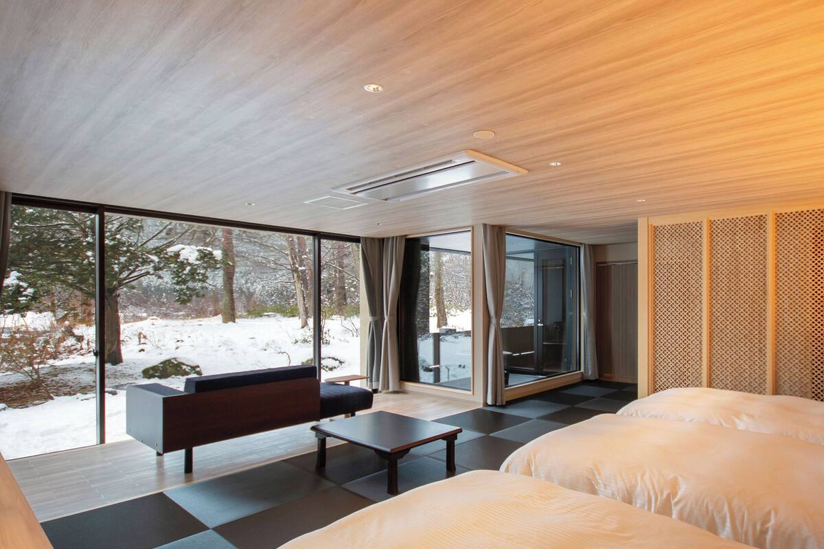 Una moderna camera d'albergo con un'ampia porta scorrevole in vetro che dà su uno spazio esterno innevato, un divano, un letto e un tavolino.