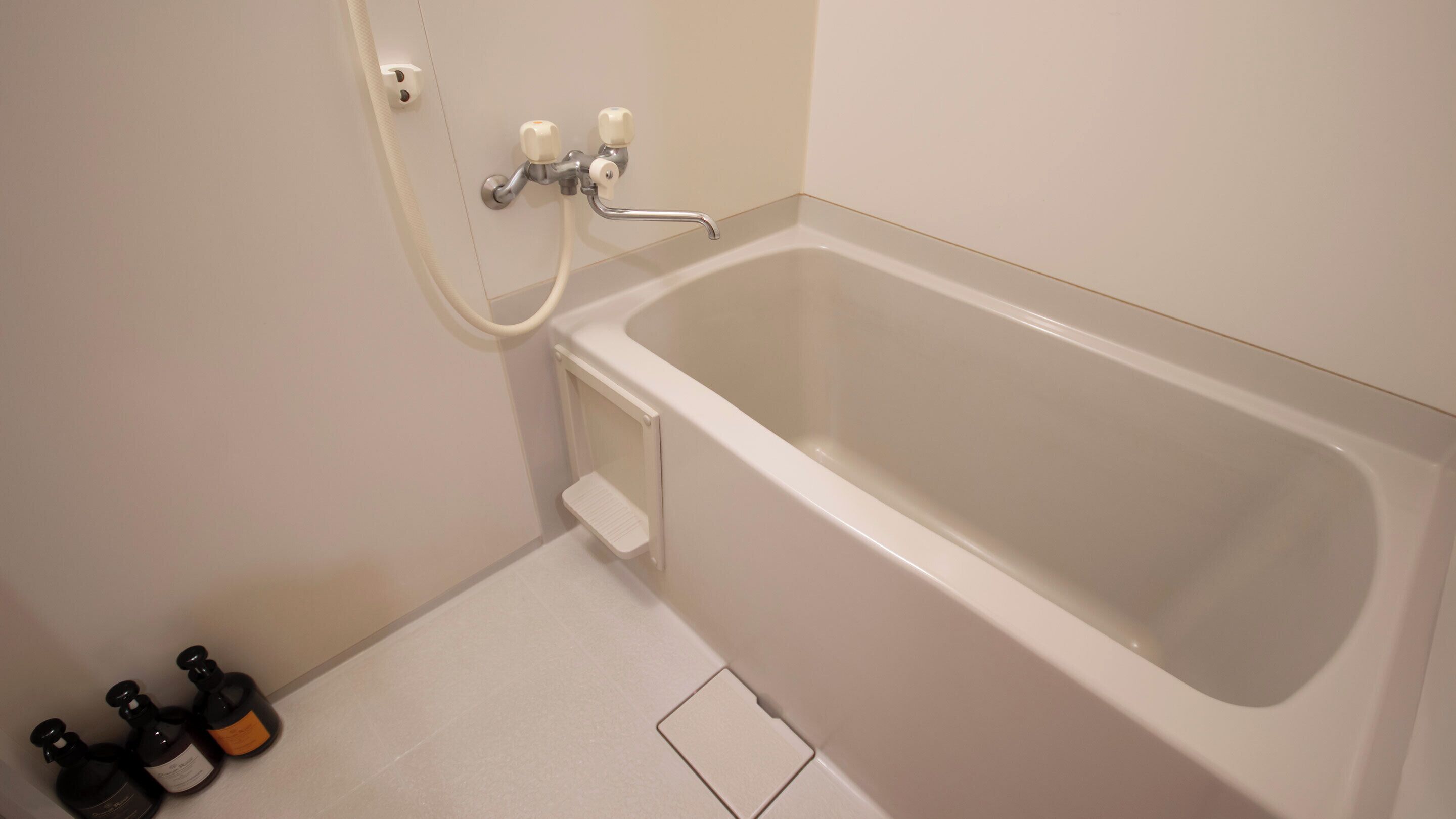 A small, clean bathroom with a white bathtub, a showerhead, and a toilet. There are three bottles of toiletries on the floor.