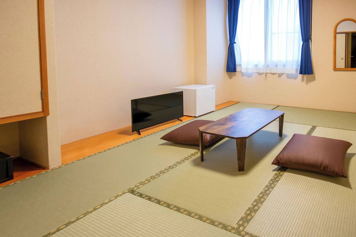 Una stanza con pavimento in tatami, un tavolino basso, due cuscini, un televisore e un elettrodomestico bianco.