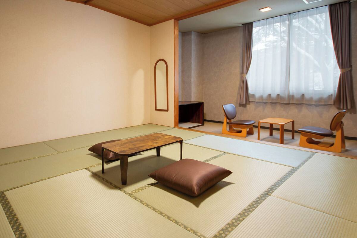 Una stanza tradizionale con tatami, un tavolino basso, una panca imbottita e due sedie di legno.