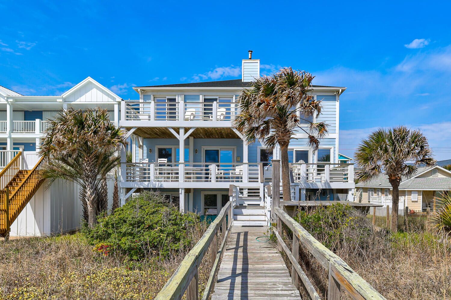 A two-story beachfront house with a wooden deck leading to the entrance, surrounded by palm trees and beach vegetation.