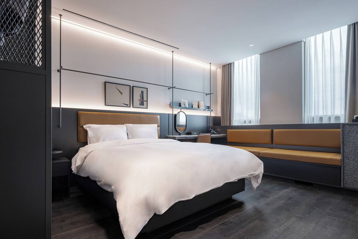 Una habitación de hotel moderna con una cama grande, mesitas de noche, un escritorio, un sofá y un gran ventanal con cortinas.