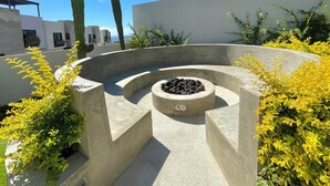 A circular concrete fire pit surrounded by yellow flowering plants, with a clear blue sky and a modern building in the background. - Marea Sabina Luxury Ocean View & Amenities (Cabo San Lucas)