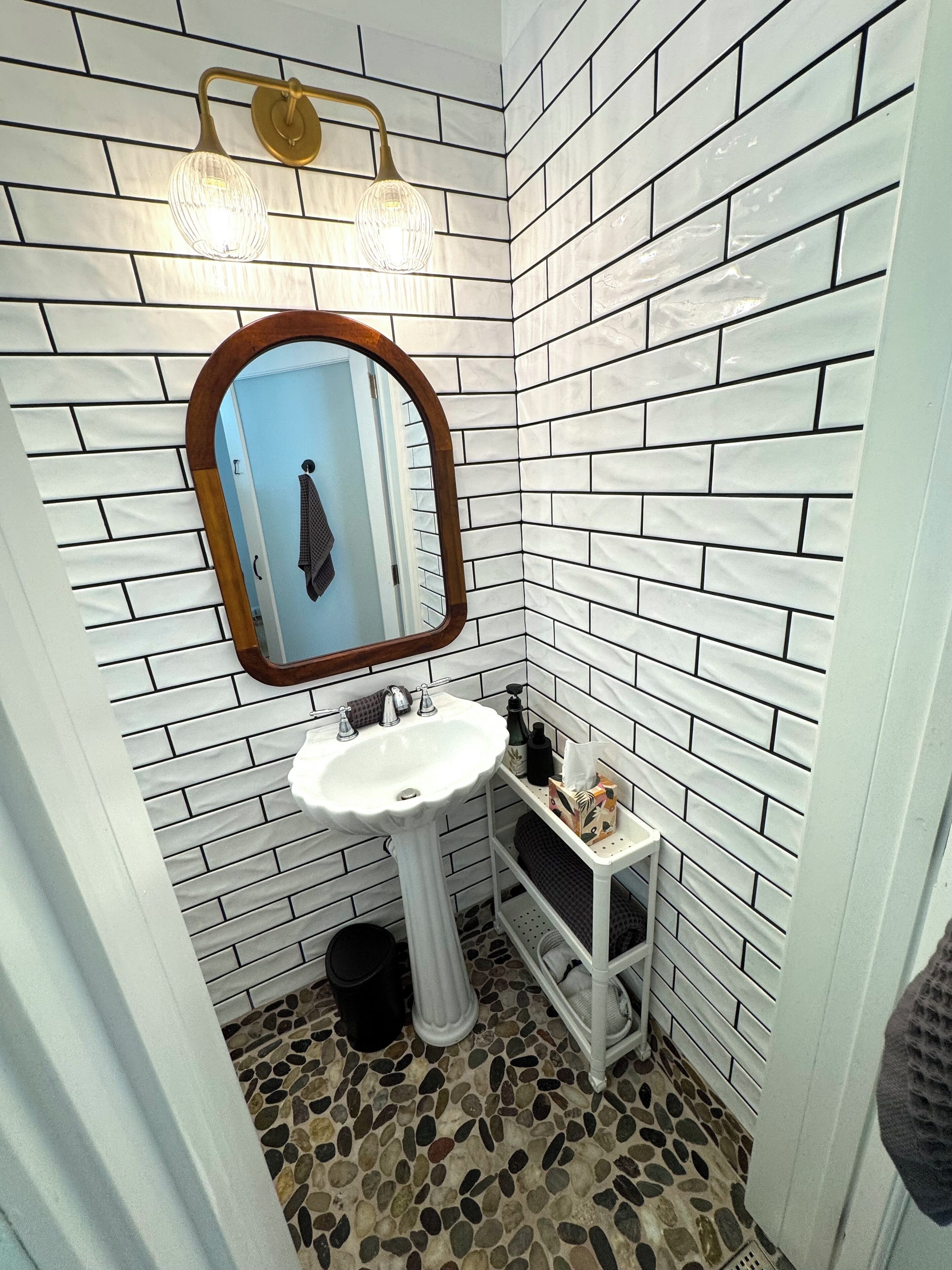 Une salle de bain avec des murs recouverts de carreaux de céramique blancs rectangulaires, un lavabo sur pied blanc, un miroir avec un cadre en bois, une petite étagère contenant des articles de toilette et un sol en mosaïque.