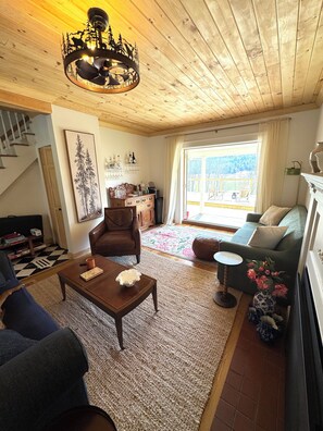 Ein gemütliches Wohnzimmer mit Holzbalkendecke, einer Kandelaberleuchte, einem Sofa, einem Couchtisch und Blick auf einen Balkon.
