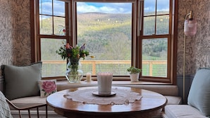 A cozy room with a round wooden table, a bench, and a window with a view of a forested landscape. - NEW! Farmhouse Mountain View’s,FireplaceGrill, Pet-Friendly, Soaking Tub (Windham)
