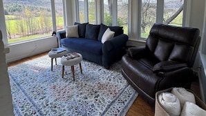 A living room with a blue sofa, a leather chair, a patterned rug, and large windows with a view of a green landscape. - NEW! Farmhouse Mountain View’s,FireplaceGrill, Pet-Friendly, Soaking Tub (Windham)