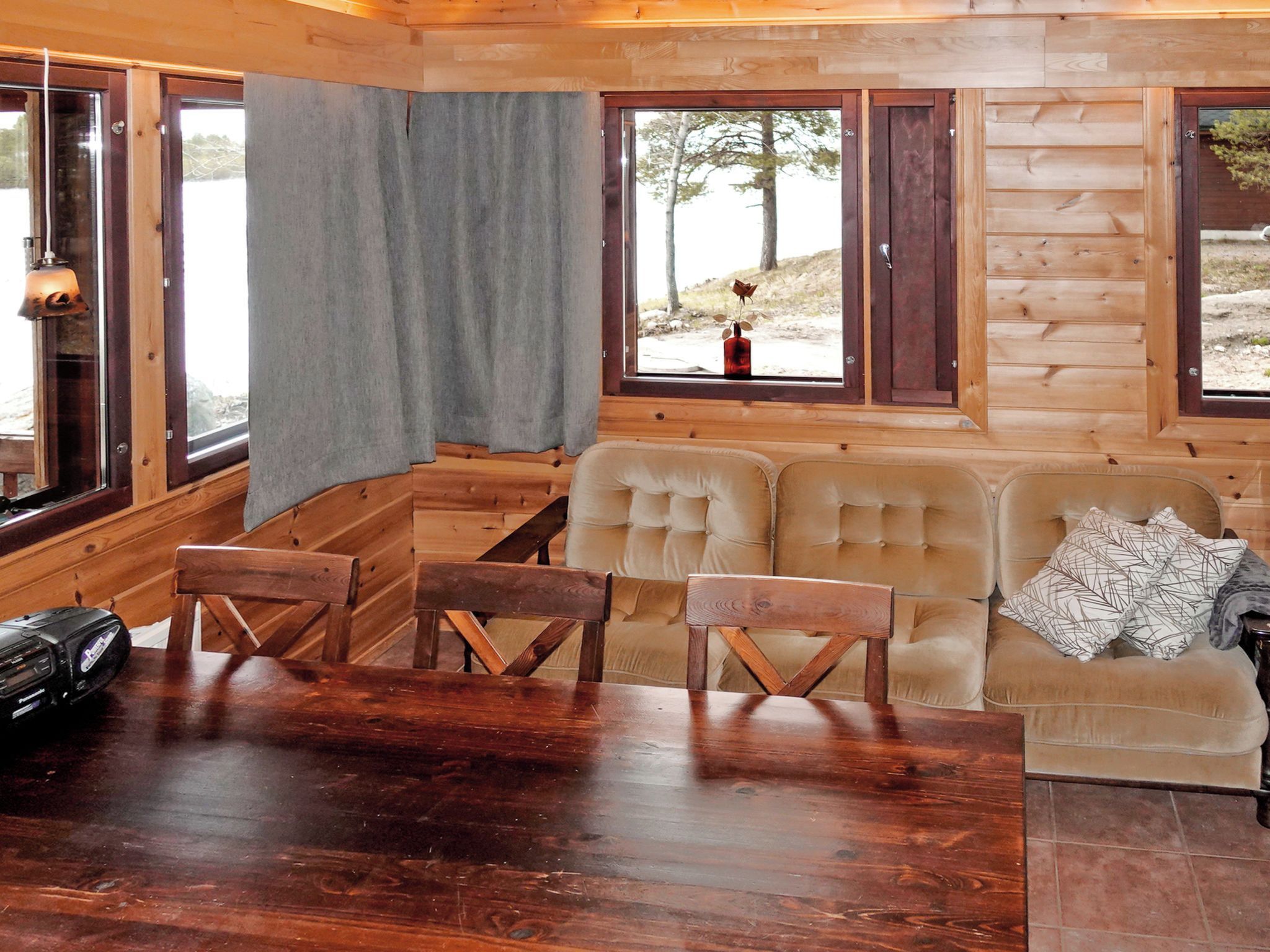 Interior de una cabaña de madera con una mesa de comedor, sillas, un sofá y ventanas amplias.