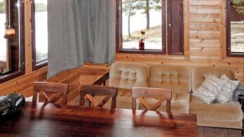 Interior de una cabaña de madera con una mesa de comedor, sillas, un sofá y ventanas amplias.