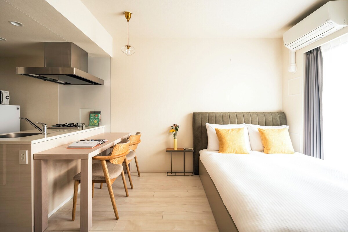 A modern hotel room with a kitchenette, a bed with two pillows, a small bedside table with a vase, and a window with curtains.