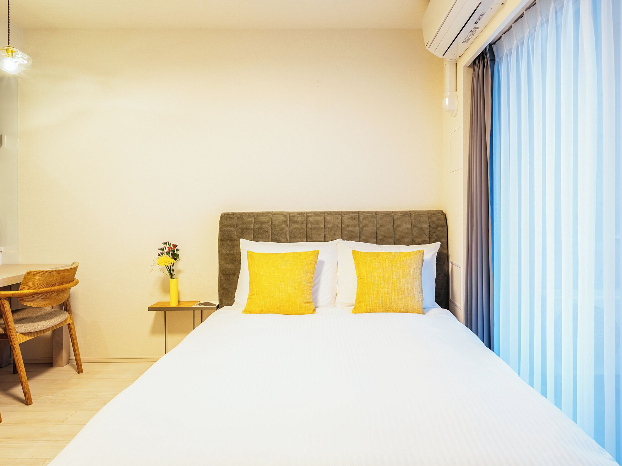 A hotel room with a bed, a chair, a small table with a vase, and a window with curtains.
