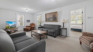 A living room with a sofa, armchair, coffee table, and a television. There are windows with blinds, a painting on the wall, and a ceiling fan.