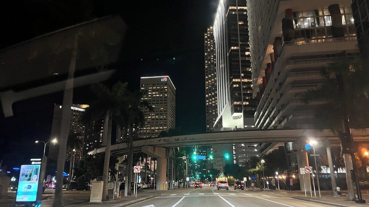 A nighttime cityscape with high-rise buildings, a pedestrian overpass, and streetlights.
