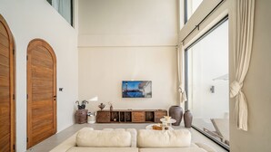 A modern living room with a large window, a sofa, a cabinet with decorative items, and a painting on the wall. - Villa Vista al Mar (Uluwatu)