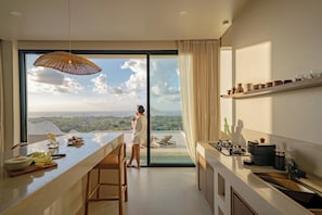 A modern kitchen with a large island, a dining area, and a view of a pool and landscape. - Villa Vista al Mar (Uluwatu)