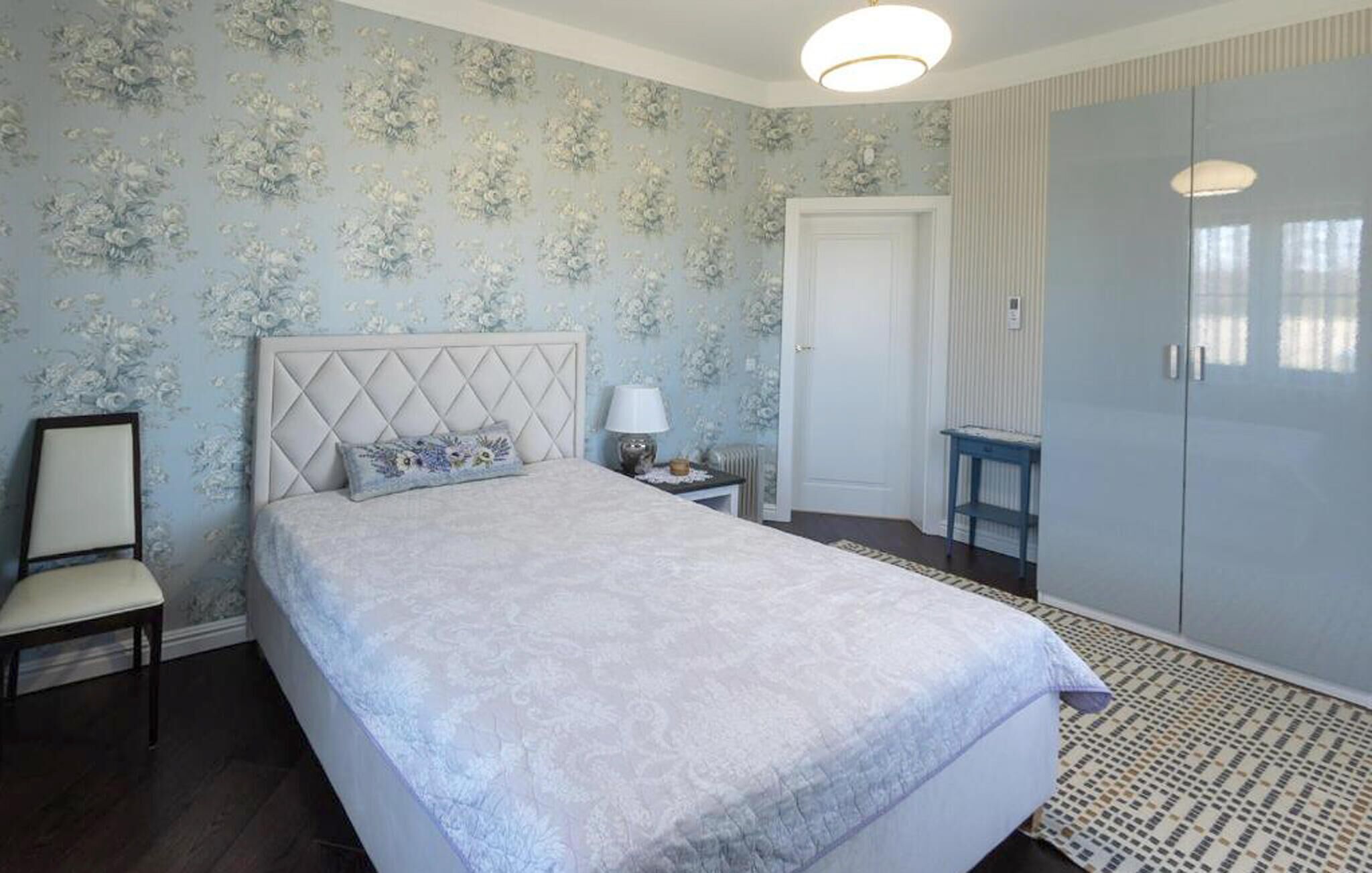A bedroom with a bed, a bedside table with a lamp, a chair, a wardrobe, and a patterned rug.