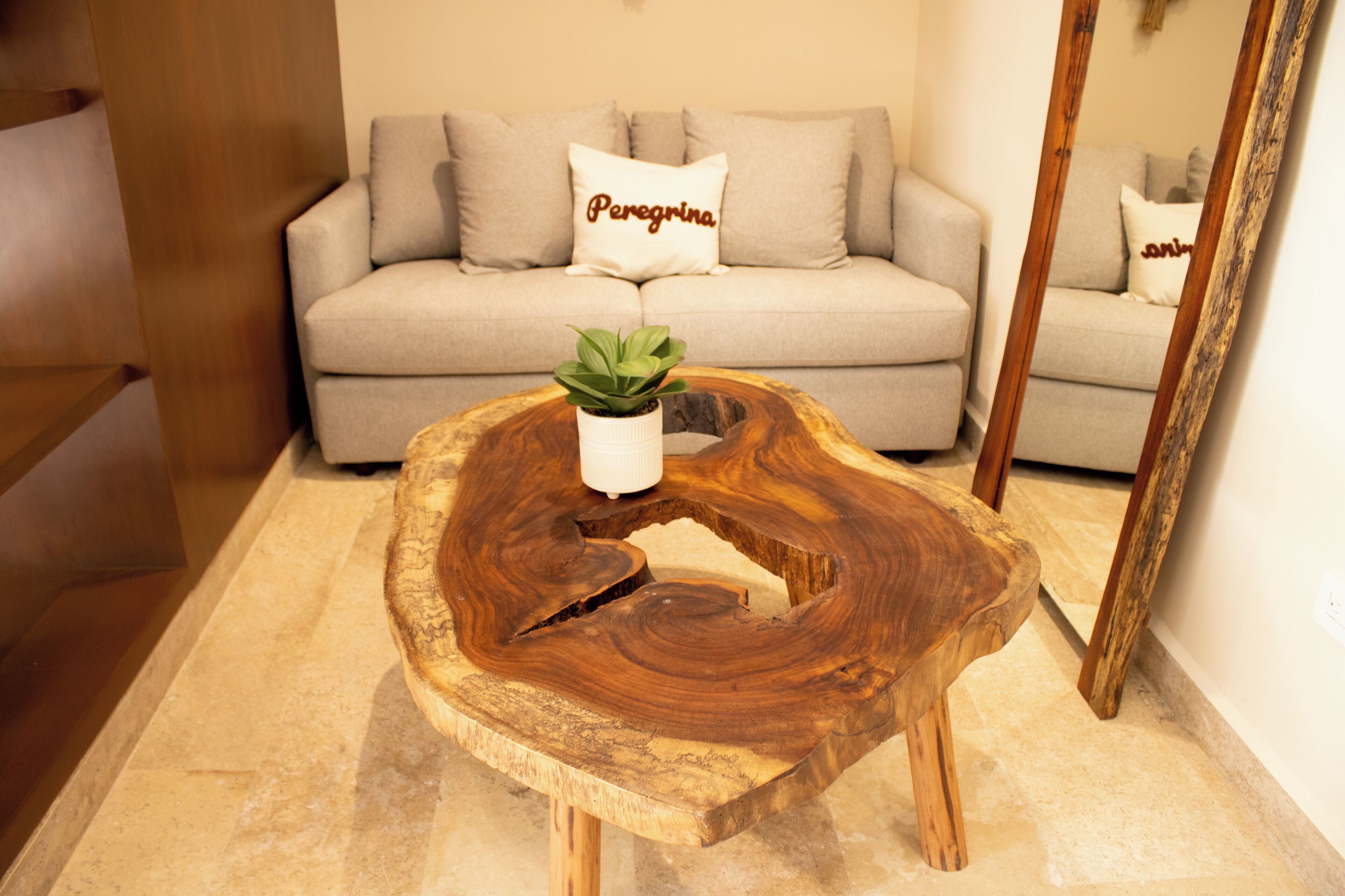 Une table basse en bois avec une plante, un canapé beige avec un coussin et un miroir dans une pièce au plancher en bois.