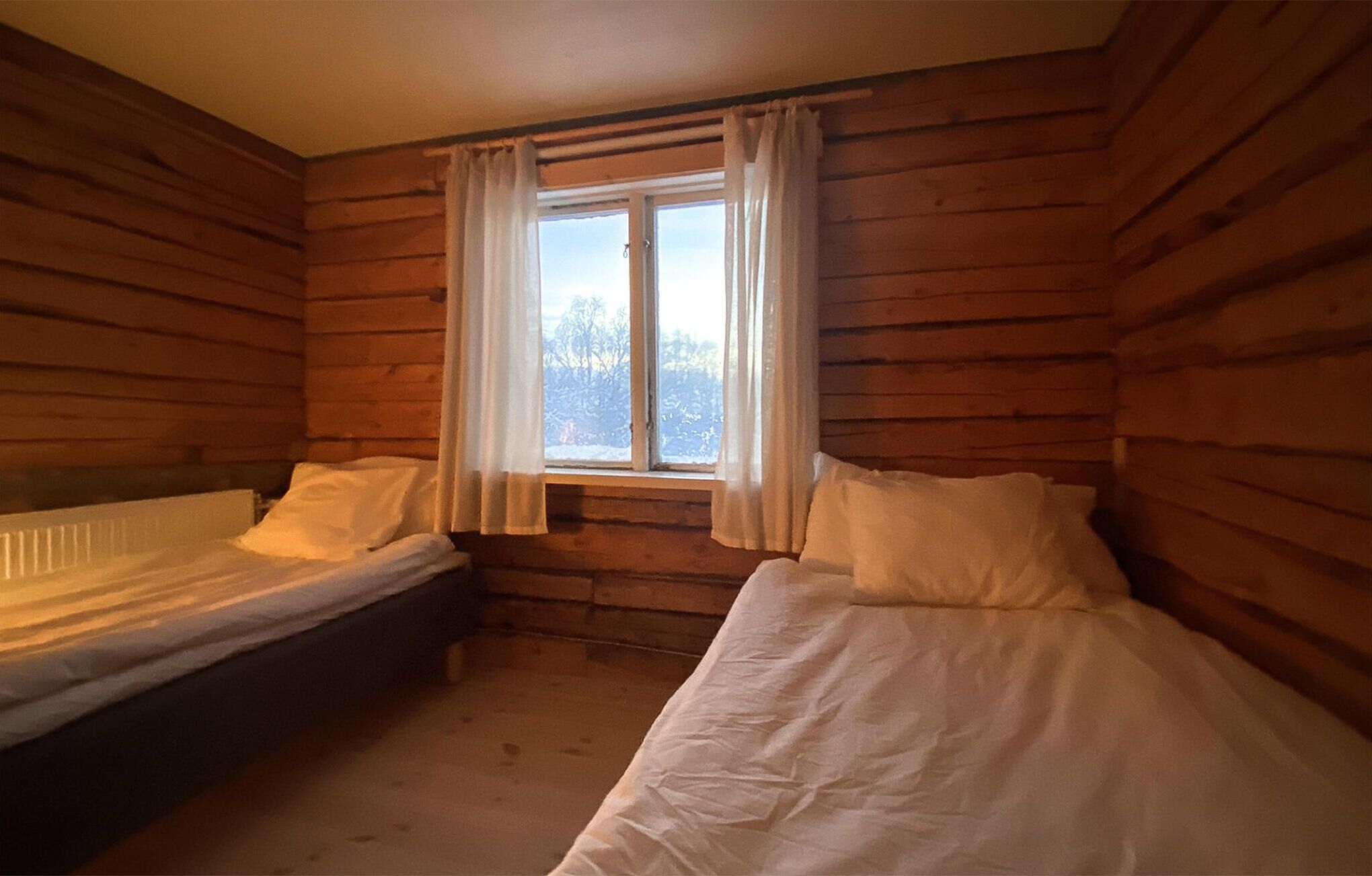 Une chambre de cabane avec deux lits, des murs en bois, une fenêtre avec des rideaux et une vue sur les arbres à l’extérieur.