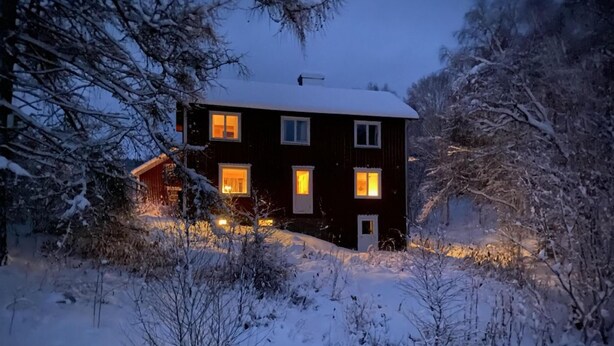 A two-story house with multiple lit windows, surrounded by snow-covered trees and ground.