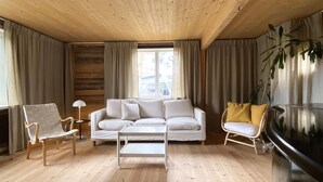 A cozy living room with a wooden ceiling, a white sofa, a small white table, a wooden armchair, a yellow armchair, and a TV.