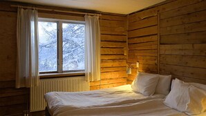 A wooden cabin room with a bed, a window with curtains, and a wall-mounted lamp.