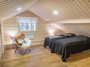 Un acogedor dormitorio en el ático con techo inclinado, ventana con vista a árboles, piso de madera, una silla con cojín mullido y dos camas con ropa de cama oscura.