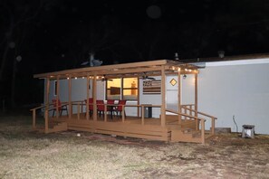 Un espacio de estar al aire libre techado con una mesa y sillas, conectado a un edificio mediante una terraza de madera.