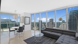 A modern living room with a large window, a gray sofa, a glass coffee table, and a dining area with chairs and a table. - Luxury Penthouse Brickell Water & City Views (Miami)