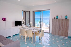 A modern dining area with a table set for four, a television on a white cabinet, and a view of the sea through large glass doors. - Villa Fortunata - Small Apartment Left, Terracina, Italy (Terracina)