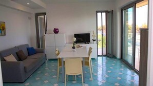 A modern living room with a sofa, a dining table with chairs, a flat-screen TV, and a large window with a view of the outdoors. - Villa Fortunata - Small Apartment Left, Terracina, Italy (Terracina)