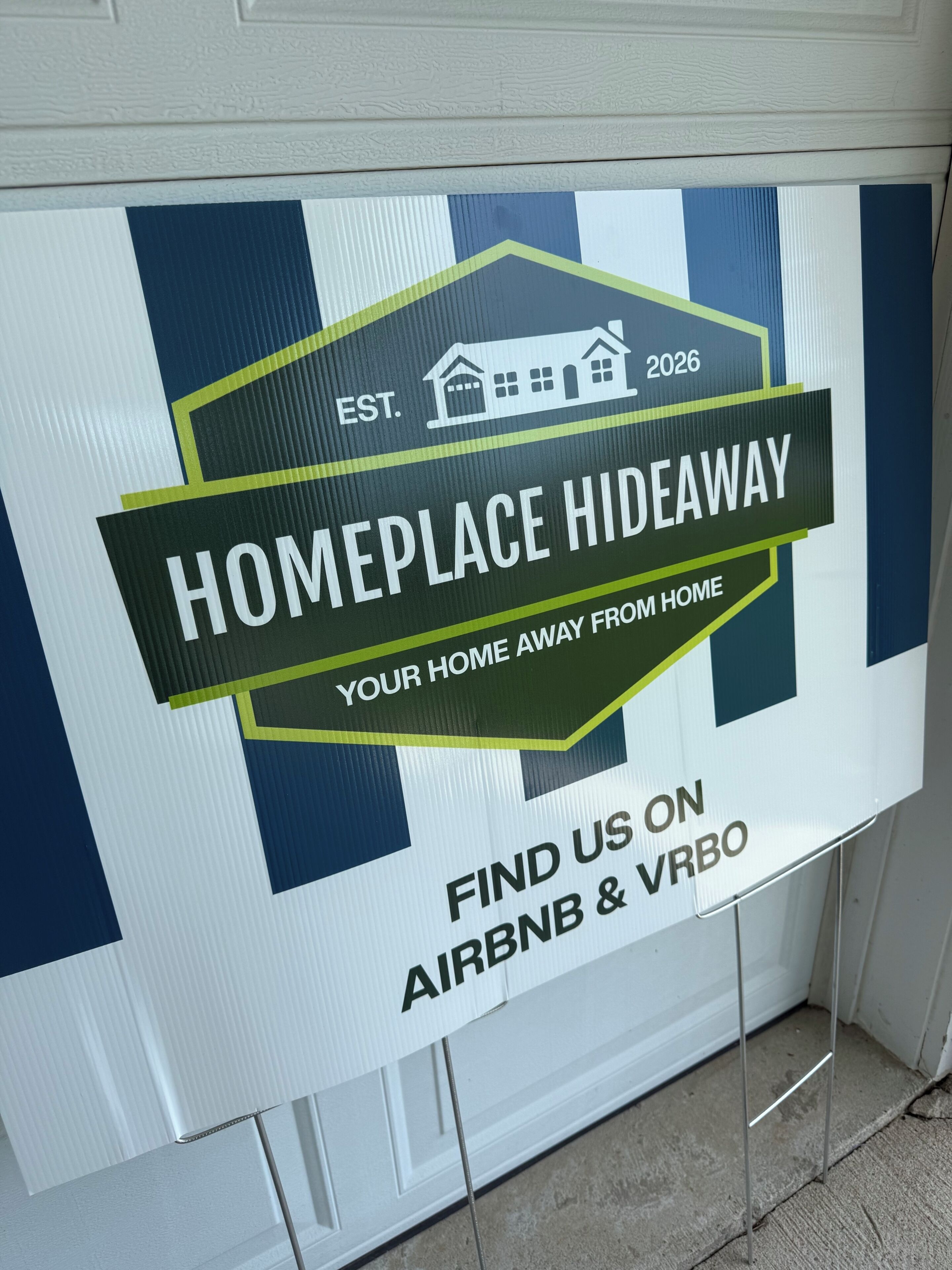 A sign advertising a 'HOMEPLACE HIDEAWAY' with a house icon, establishment year 2026, and mentions Airbnb & VRBO.