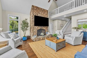 A living room with a stone fireplace, a TV mounted above it, a staircase to an upper level, and a view of greenery outside.