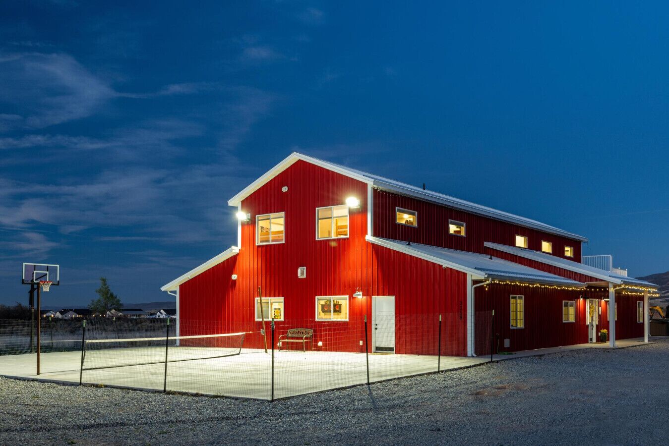 A red barn with a basketball court in front, a covered area with string lights, and a clear sky.
