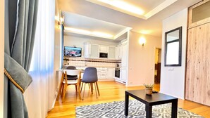Un saló modern amb sòl de fusta, una catifa amb dibuixos, una zona de menjador amb cadires, una cuina amb armaris blancs i un televisor penjat a la paret.