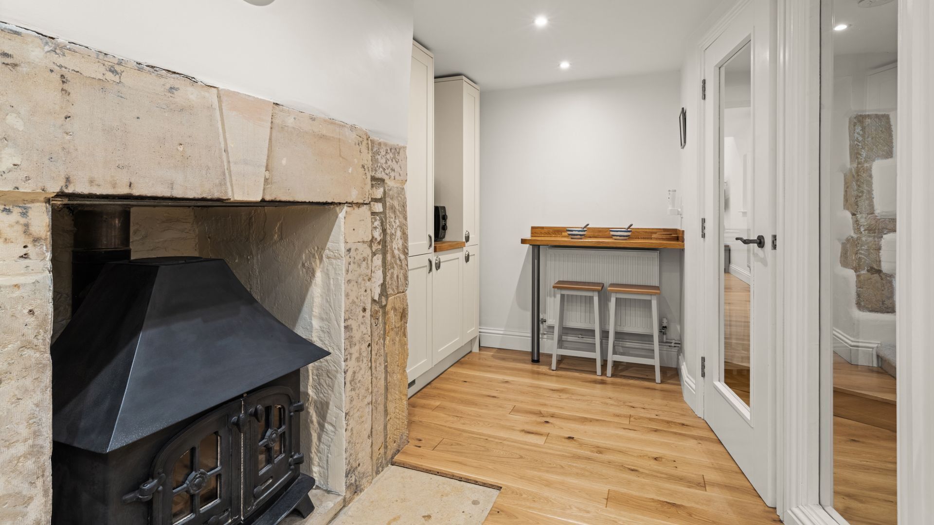 A kitchen with a stone fireplace, wooden flooring, and white cabinetry.