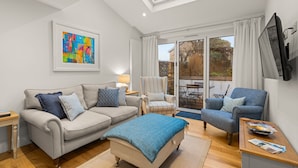 Un salon moderne avec un canapé, un fauteuil et un pouf, un tableau accroché au mur et un téléviseur fixé au mur.