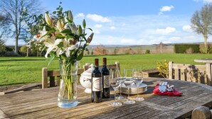 Ein Holztisch im Freien mit Weinflaschen, Gläsern und einem Blumenstrauß.