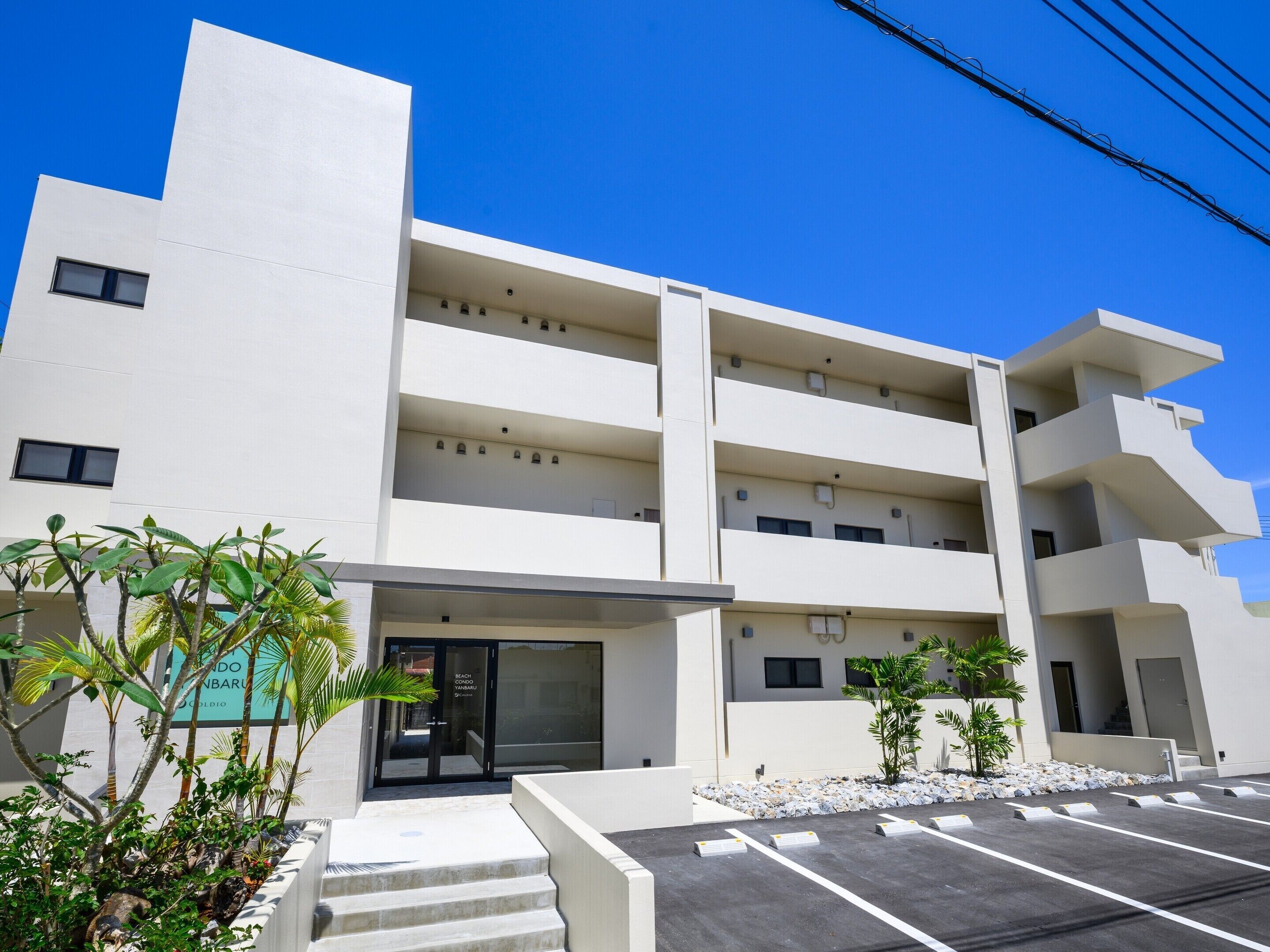 A multi-story building with a modern design, featuring a parking area with marked spaces and a staircase leading to the entrance.
