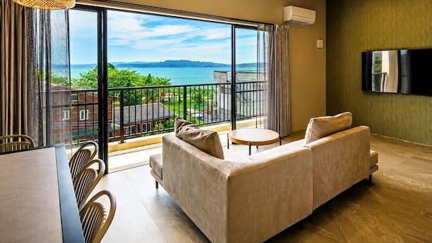 A modern living room with a sofa, a dining table with chairs, a TV mounted on the wall, and a balcony with a view of the sea. - Yanbaru Pool Condo Yagaji by COLDIO (Nago)