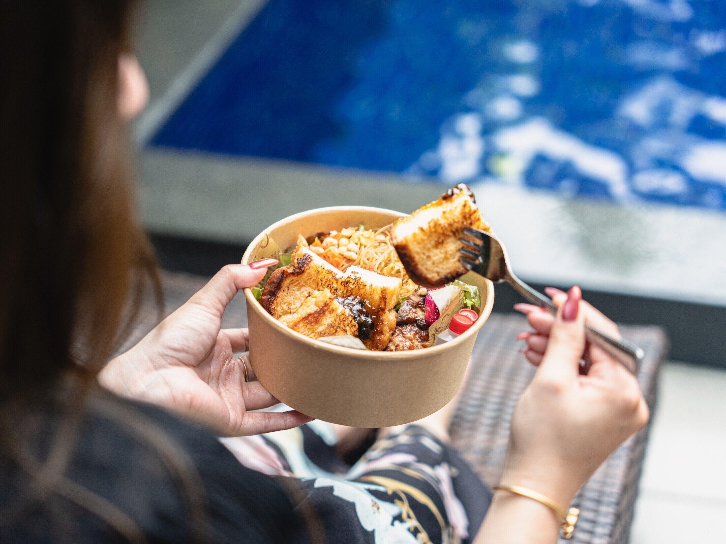 A person holding a bowl with a meal, sitting near a pool.