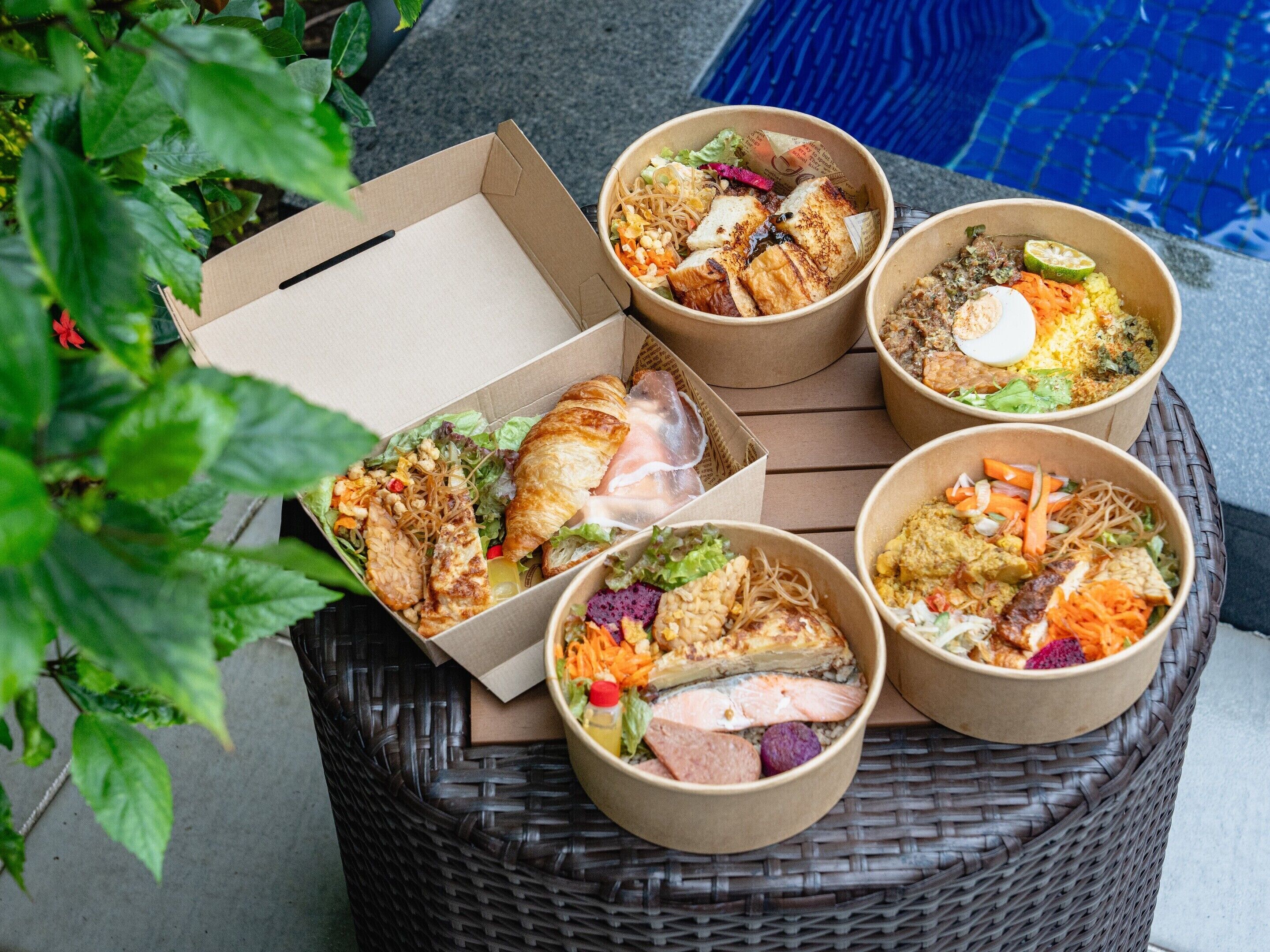 A variety of food items in paper containers, including salads, noodles, and grilled meats, arranged on a wicker table near a pool.