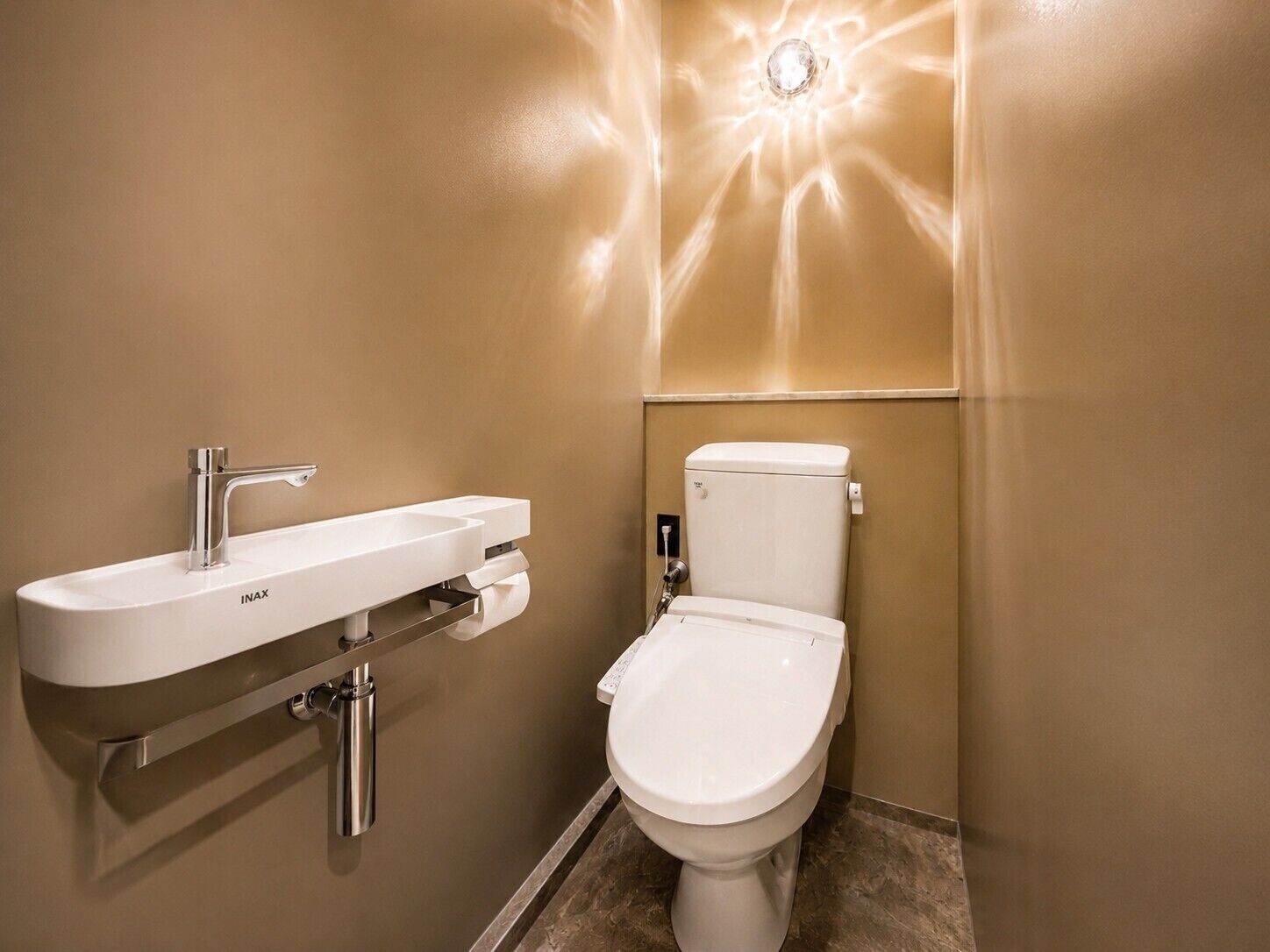 A bathroom with a white INAX sink, a toilet, and a wall-mounted light fixture.